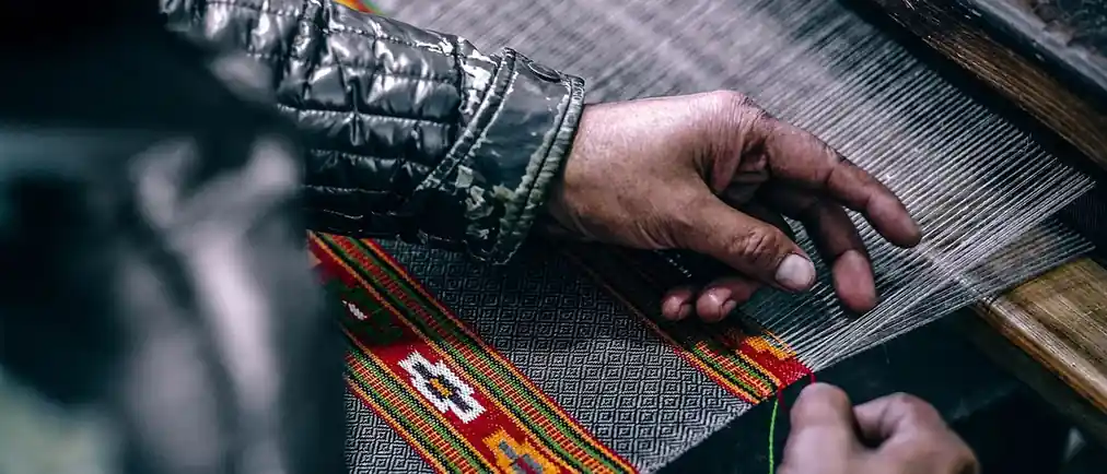 Two hands guiding red and gold patterned cloth through a loom