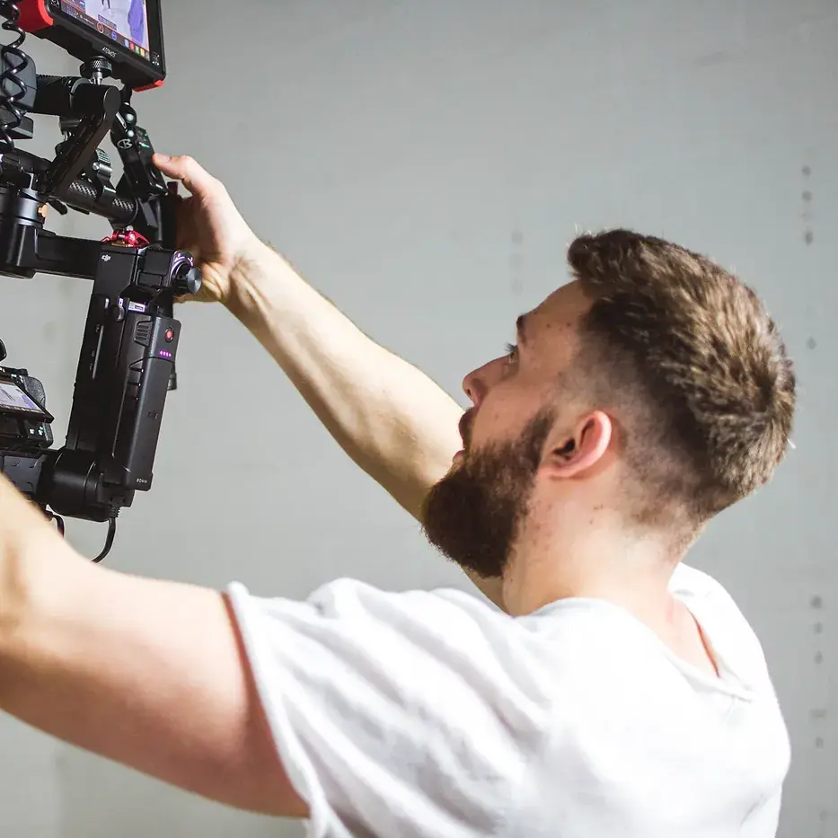 A camera operator raising a hand-held rig to eye level against a plain wall