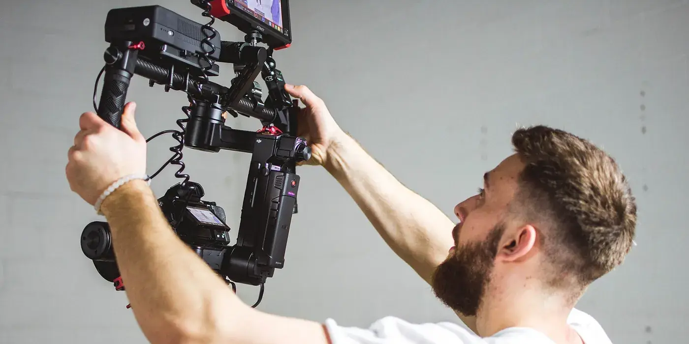 A camera operator in a white T-shirt lifting a hand-held camera rig with both hands